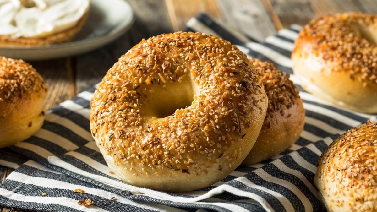 Draufsicht: Auf einem Holzliegt liegt ein schwarz-weiß gestreiftes Küchenhandtuch. Auf dem liegen mehrere Bagels, die mit Sesam bestreut wurden.