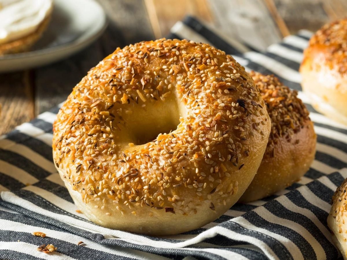 Draufsicht: Auf einem Holzliegt liegt ein schwarz-weiß gestreiftes Küchenhandtuch. Auf dem liegen mehrere Bagels, die mit Sesam bestreut wurden.