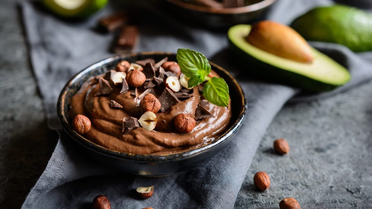 Eine schwarze Schale mit dem Avocado-Schokopudding auf grauem Untergrund. Im Hintergrund unscharf eine Avocado und ein paar Nüssen.