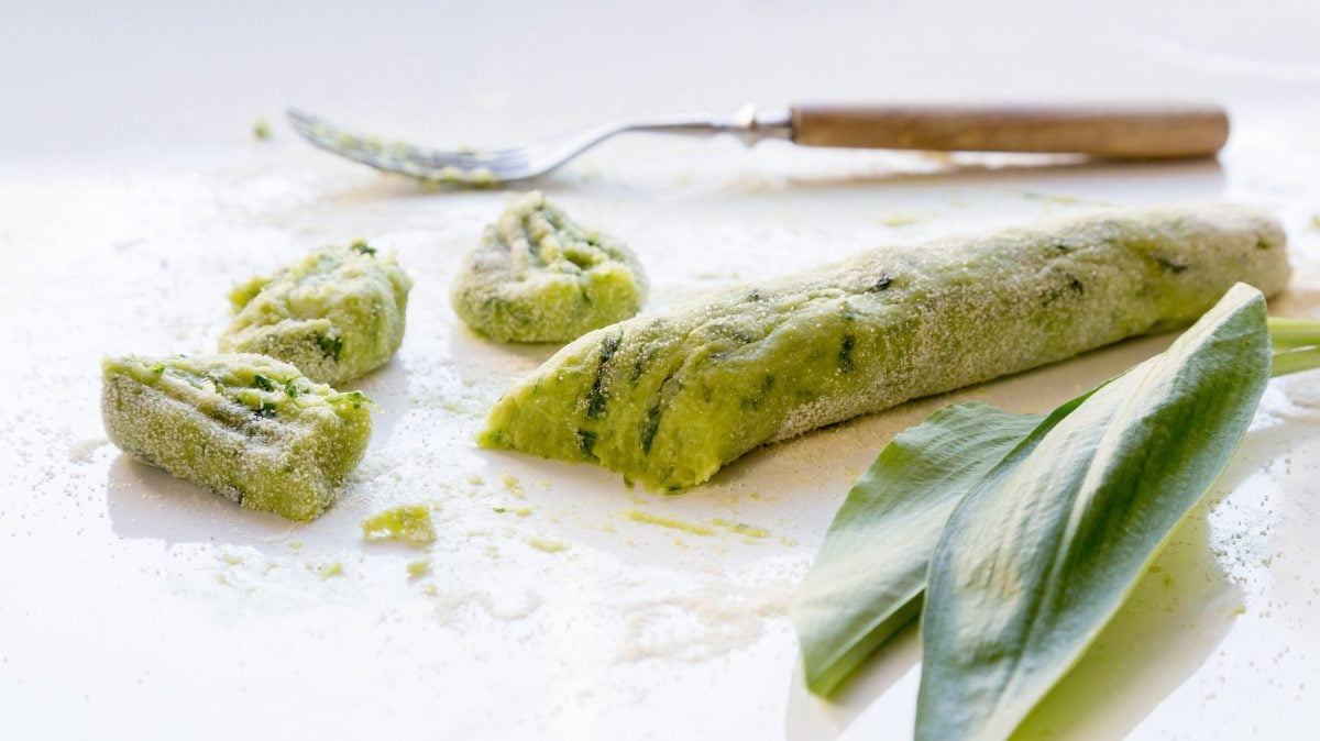 Eine Rolle Teig für Bärlauch-Gnocchi, ein paar Gnocchi sind schon abgeschnitten, daneben frischer Bärlauch und eine Gabel.