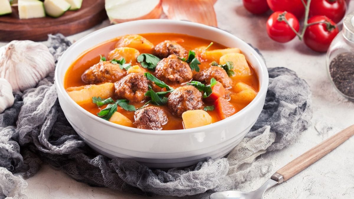Eine weiße Schale mit dem Bauerntopf auf einem hellen Tisch mit Tomaten, Zucchini und Zwiebel.