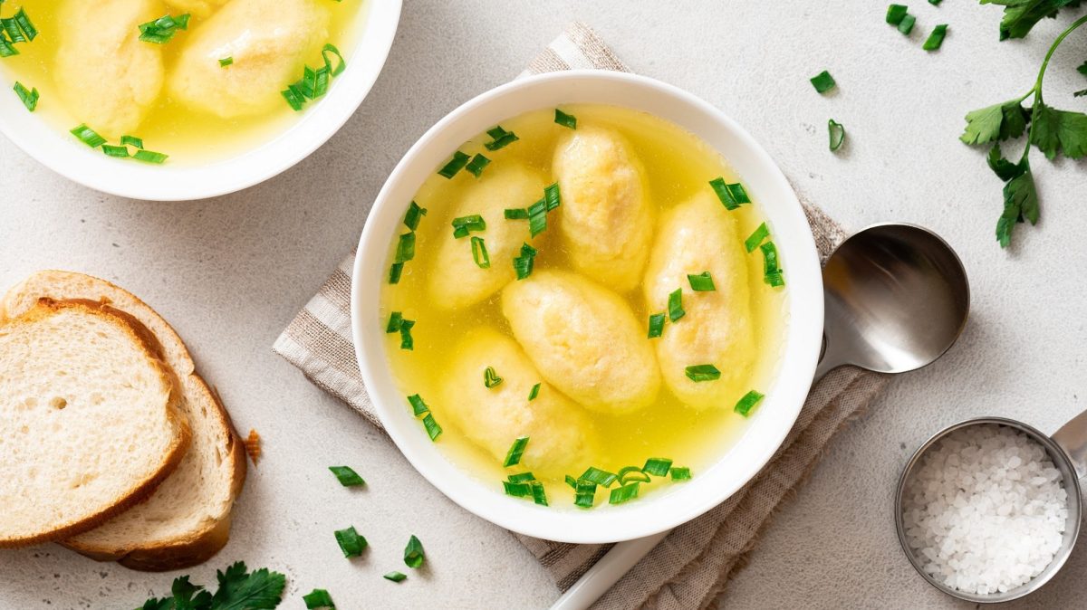 Draufsicht: zwei Schüsseln Bayerische Grießnockerlsuppe. Drumherum stehen ein Salzgefäß, Löffel, Brotscheiben und frische Kräuter.