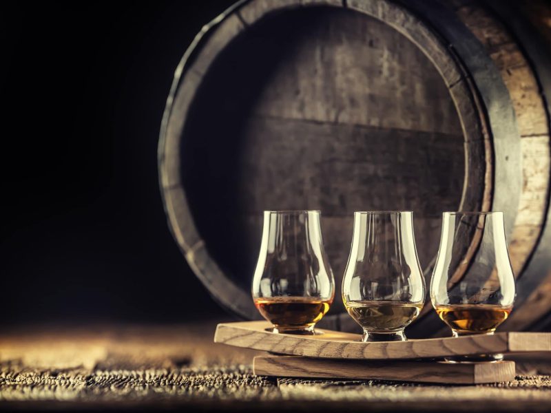 Bourbon oder Scotch? Welcher Whisky für Anfänger? Drei Gläser Whisky auf Holz vor einem Holzfass. Frontalansicht.