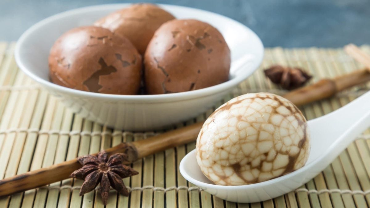 Chinese Tea Eggs chinesische Tee-Eier mit Schale in einer weißen Schüssel auf Bast. Auf weißem Löffel ein geschältes. Frontalansicht.