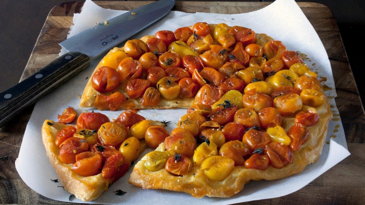 Angeschnittene Cocktailtomaten-Tarte-Tatin auf Backpapier auf dunklem Untergrund. Daneben ein Kuchenmesser. Draufsicht.
