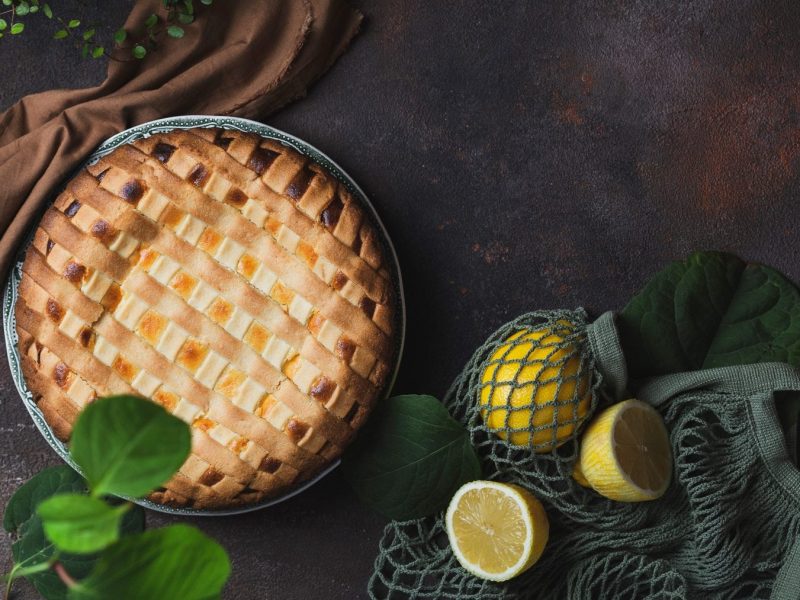 Crostata al limone in Springform auf dunklem Untergrund. Im Bild verteilt Zutaten.