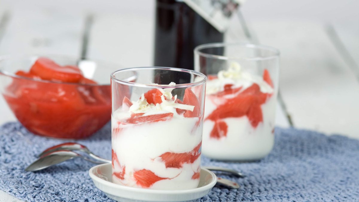 Frontalsicht: Zwei Gläser Rhabarber-Joghurt. Dahinter steht eine Schüssel aus Glas mit Rhabarber-Kompott.