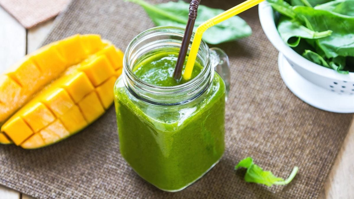 Ein Glas mit Mango-Minze-Smoothie neben frischer Mango und Spinat.