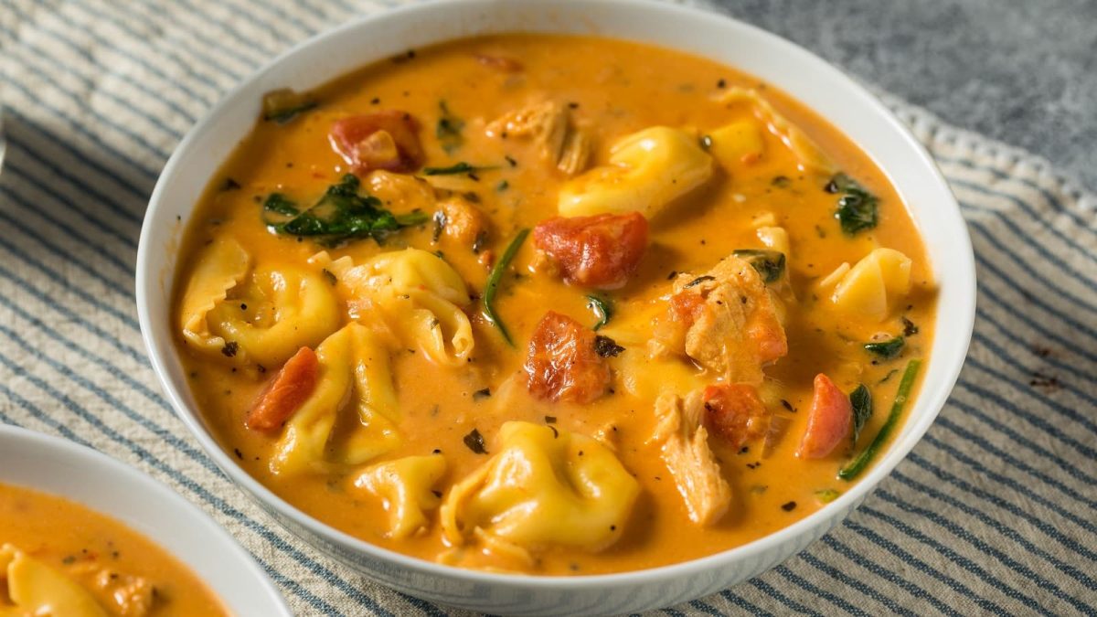 Eine weiße Schüssel Tortellini-Tomatencremesuppe auf einem hellen Tisch.