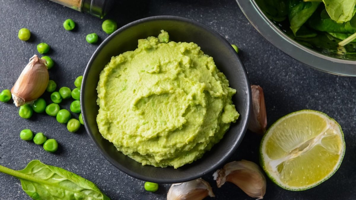 Eine schwarze Schale mit Erbsen-Hummus neben frischen Erbsen.