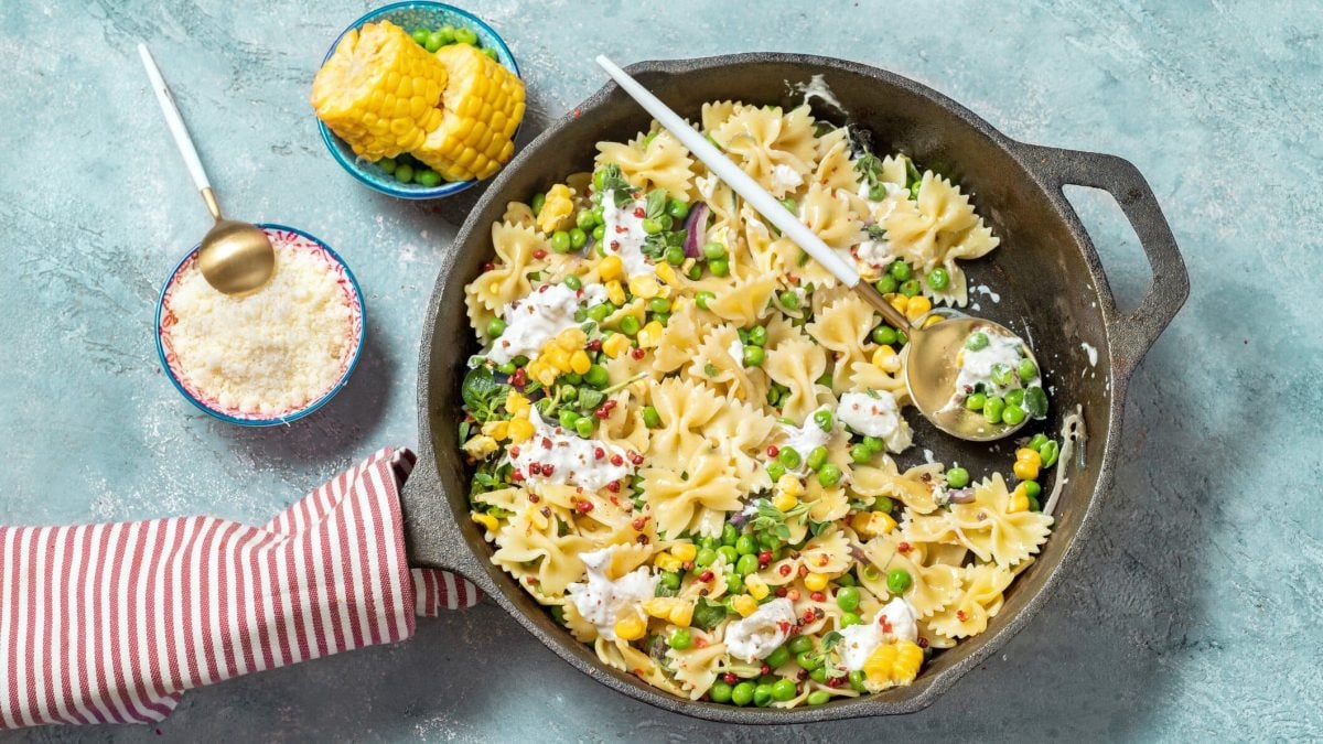 Draufsicht: vegetarische Farfalle mit Erbsen und Burrata auf einen hellblauen Untergrund. Daneben liegen eine Schüssel mit gerieben Parmesan sowie Mais