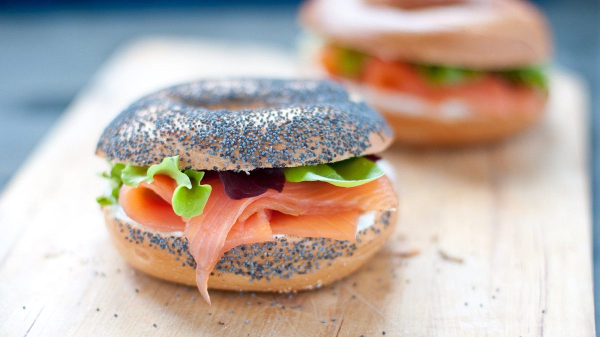 Frontalsicht: Zwei Käse-Bagel mit Lachs liegen auf einem hellen Holztisch