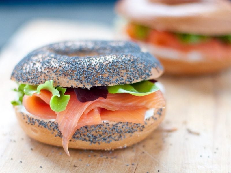 Frontalsicht: Zwei Käse-Bagel mit Lachs liegen auf einem hellen Holztisch