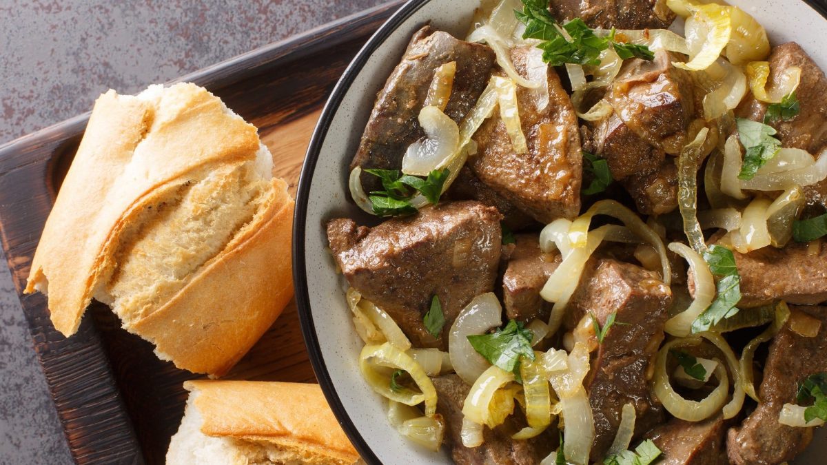 Draufsicht: EIne Schüssel mit gebratener Leber und Zwiebeln steht auf einem dunklen Tablett. Daneben liegen zwei Baguette-Scheiben.