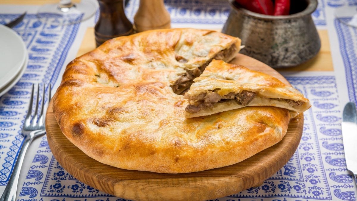 Schrägsicht von oben: Ein mit Fleisch gefülltes, rundes Fladenbrot liegt auf einem Holzbrett. Es handelt sich um das georgische Gericht Kubdari. Es wurde angeschnitten. Das Holzbrett liegt auf einem blau-weißen Tischtuch.