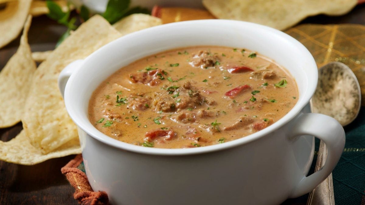 Weiße Schüssel gefüllt mit tomatiger Hackfleisch-Käse-Suppe. Daneben liegen Chips.