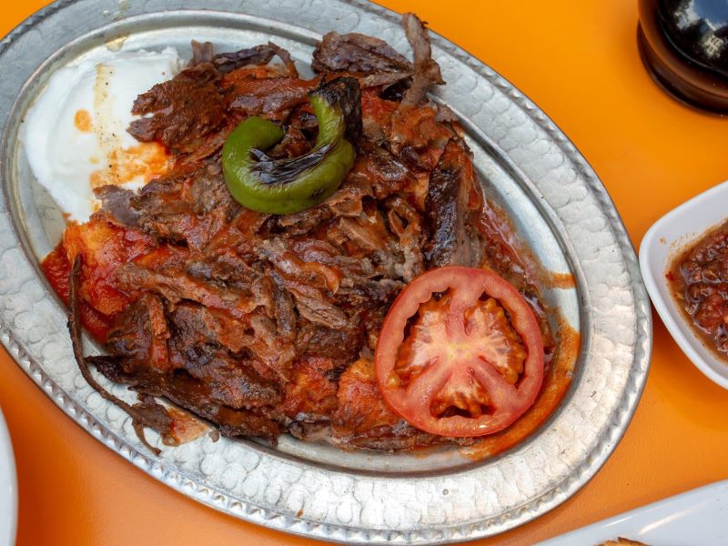 Iskender Kebab angerichtet auf Platte auf orangenen Tisch. Im Bild weitere Zutaten. Draufsicht.