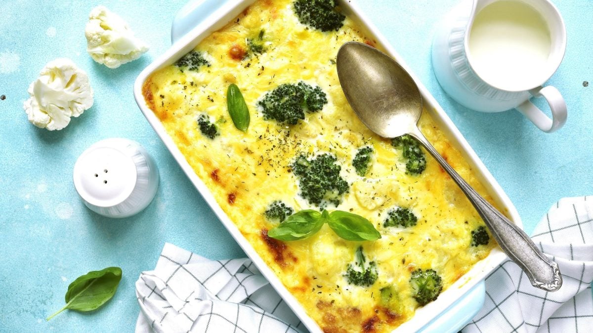 Draufsicht: Auf einem hellblauem Untergrund steht ein Brokkoli-Gratin mit Blumenkohl. Drumherum liegen Basilikumblätter und ein Kännchen mit Sahne sowie ein Salzstreuer