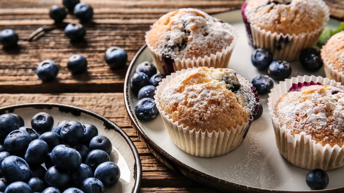 Draufsicht: Muffins mit Beeren stehen auf einem Teller, der auf einem Holztisch liegt. Daneben ist ein kleiner Teller mit Blaubeeren. Auch auf den Tisch liegen noch frische Beeren.