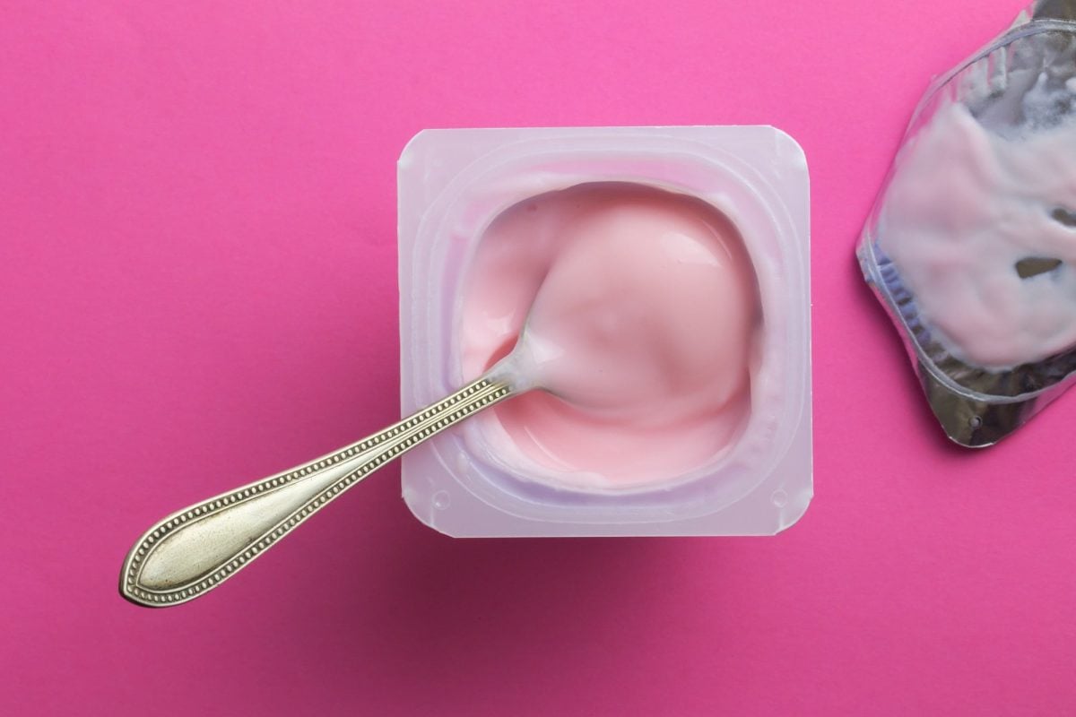 Draufsicht: Auf einem pinken Untergrund steht ein geöffneter Joghurtbecher. In dem Joghurt steckt ein Löffel. Neben dem Joghurt lieget der Joghurtdeckel.