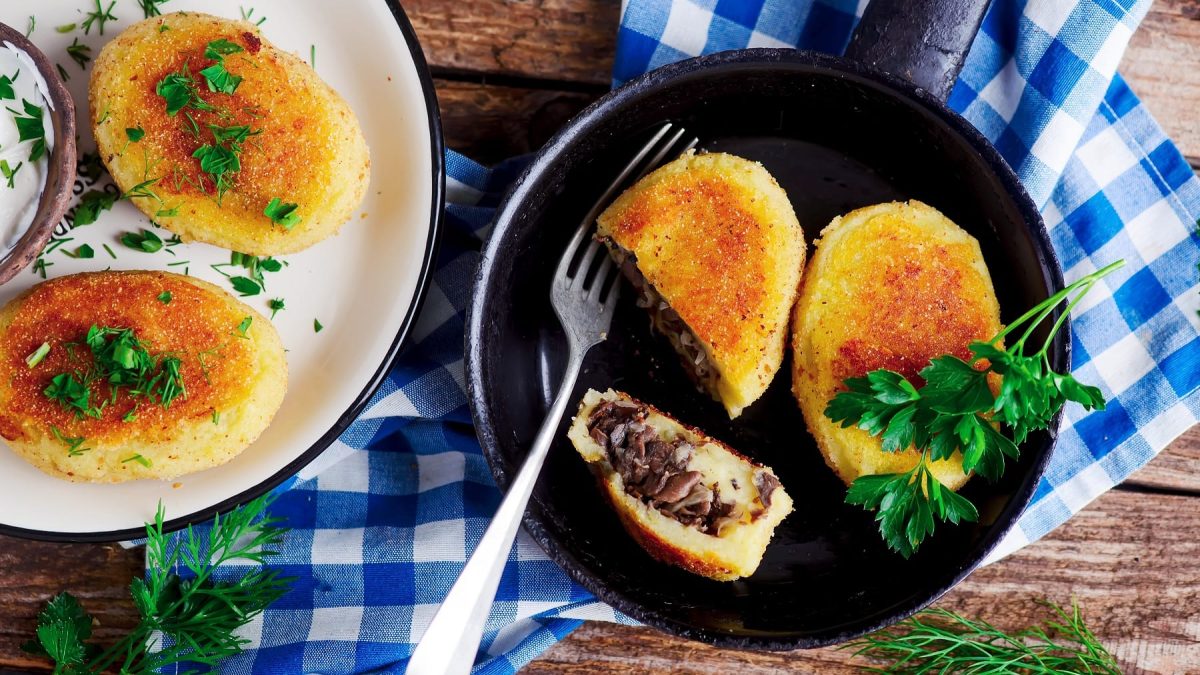 Eine Pfanne mit den Kartoffeltalern auf einem rustikalen Holztisch mit blau-weiß-karierter Tischdecke und Kräutern.