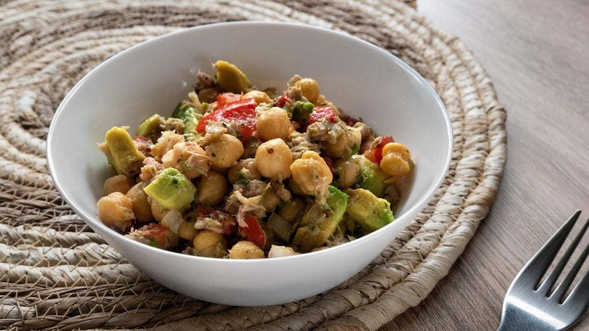 Kichererbsensalat mit Thunfisch und Avocado in weißer Schale auf Untersetzer und Holztisch. Daneben eine Gabel. Draufsicht.