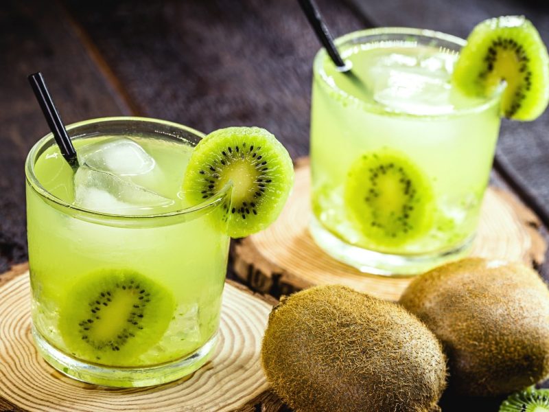 Zwei grüne Cocktails mit Kiwi auf zwei Holzscheiben und dunklem Untergrund.