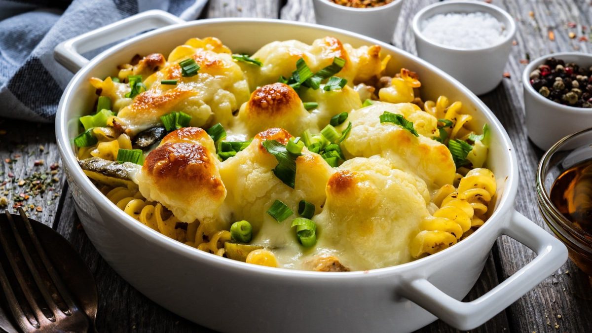 Schräge Draufsicht: Eine weiße Form mit einem Blumenkohl-Hackauflauf der mit Käse überbacken wurde. Der steht auf einem dunklen Holztisch. Daneben liegen mehrere kleine Gefäße mit Gewürzen, wie etwa Salz und Pfeffer, sowie zwei Gabeln und ein Küchentuch.