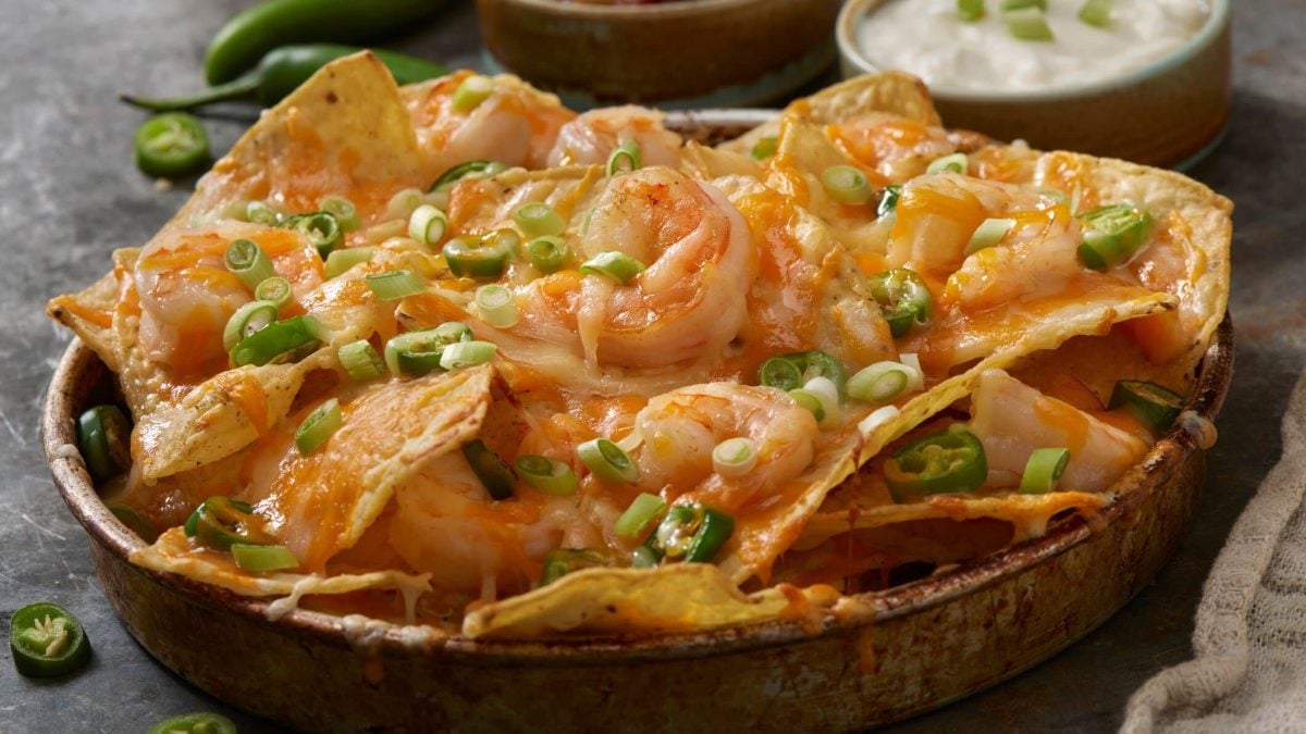 Loaded Garlic Shrimp Nachos in einer Tonform auf dunklem Untergrund. Im Hintergrund zwei Schalen mit Dip. Draufsicht.