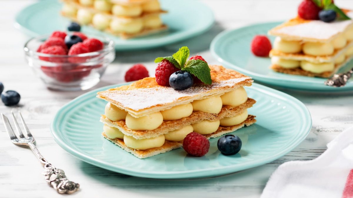 Drei blaue Teller mit dem Mille-feuille auf einem hellen Holztisch mit eine rweißen Serviette und einer silbernen Gabel.