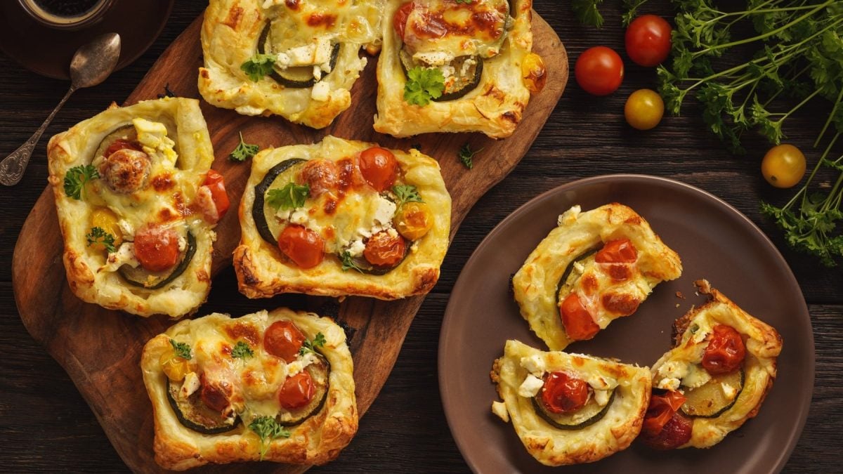 Mehrere Mini-Blätterteig-Tartes mit Zucchini, Tomaten und Feta in der Draufsicht.