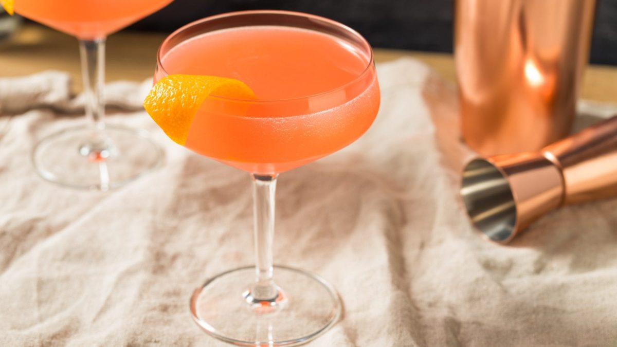 Monkey Gland Gin-Cocktail mit Absinth in Cocktailschale. Im Hintergrund Barmaß, Shaker und ein zweiter Monkey Gland angedeutet. Frontalansicht.