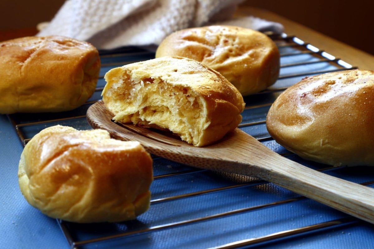 Fünf Pan de Coco auf Rost auf blauem Untergrund. Eine halbiert auf Holzkochlöffel. Draufsicht.