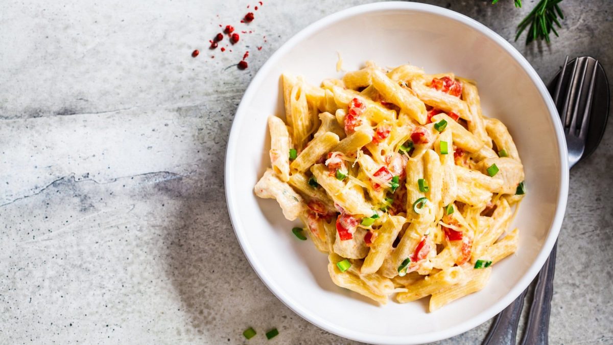 Ein Teller Pasta mit Hummus-Sauce, Frühlingszwiebeln und Paprika in der Draufsicht, daneben Besteck.