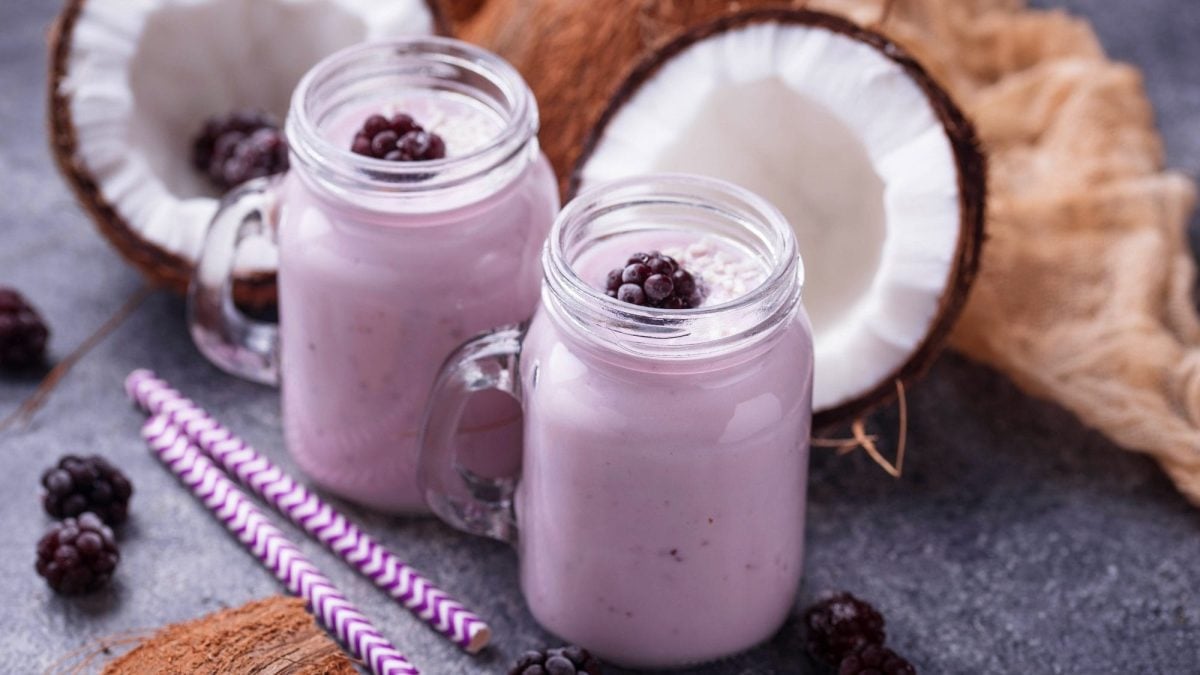 Schräge Draufsicht: Zwei Gläser Kokos-Brombeer-Shake stehen auf einem dunklen Untergund. Daneben liegen zwei geringelte Strohhalme, zwei Kokosnuss-Hälften und Brombeeren.