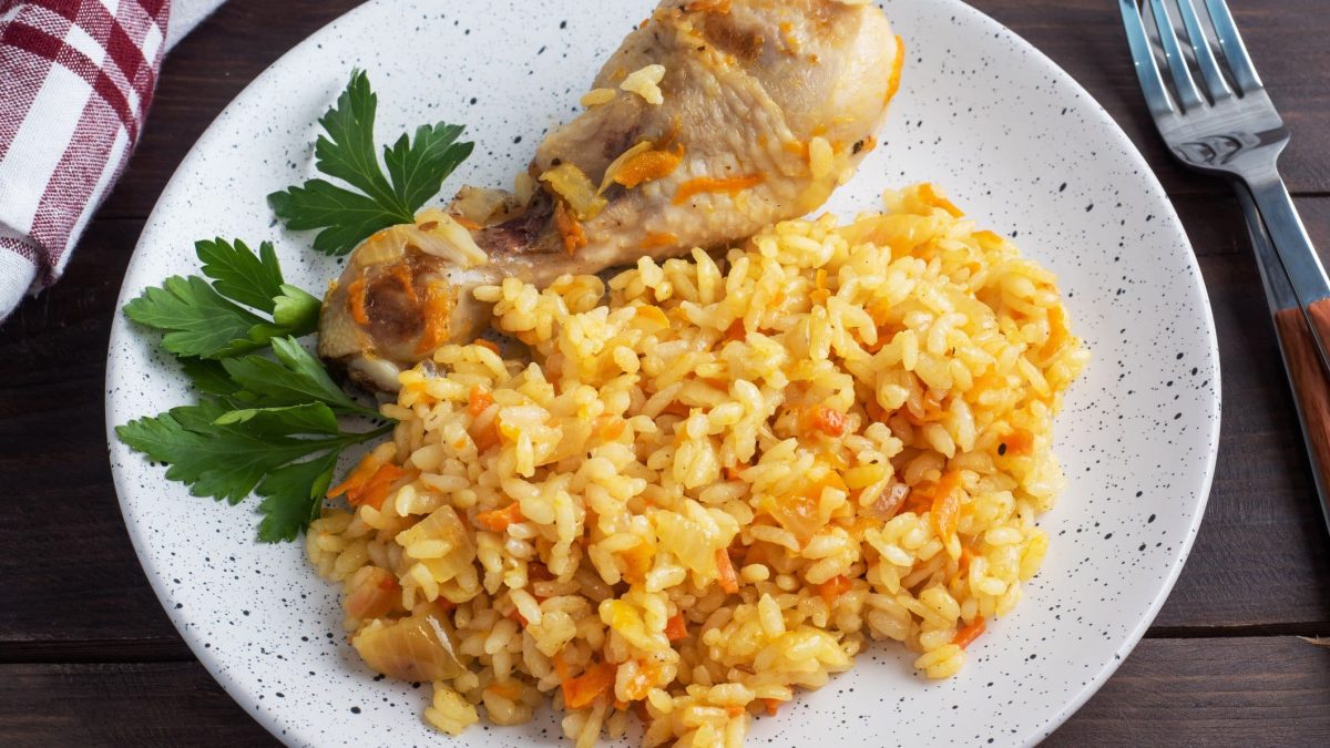 Plov mit Drumstick auf weißem Teller mit Petersilie. Daneben Gabel und Geschirrtuch. Draufsicht.