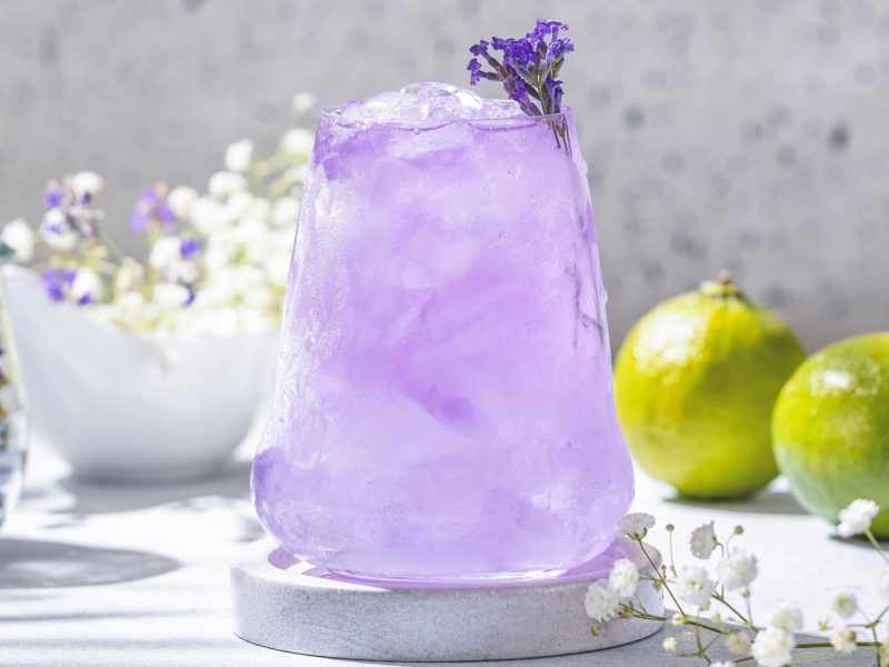 Ein Glas mit dem violetten Cocktail auf hellem Untergrund. Im Hintergrund Limetten, Anchan-Tee und lila Blüten.