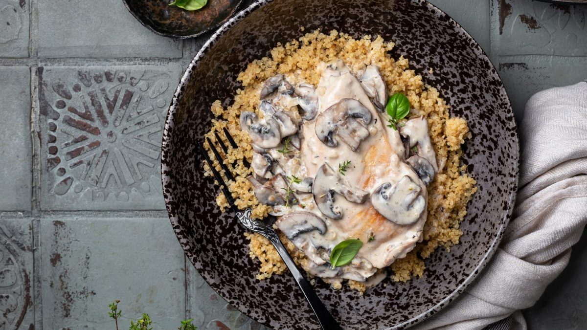 Ein schwarzer Teller mit Quinoa und dem Pilz-Rahm auf grauen Fliesen mit einer schwarzen Gabel.