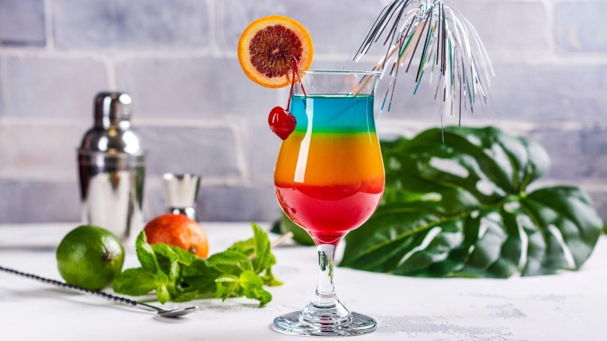 Ein Glas mit Regenbogen-Cocktail (unten rot, Mitte gelb-orange, oben blau), garniert mit Kirsche und Blutorangenscheibe vor hellem Hintergrund.
