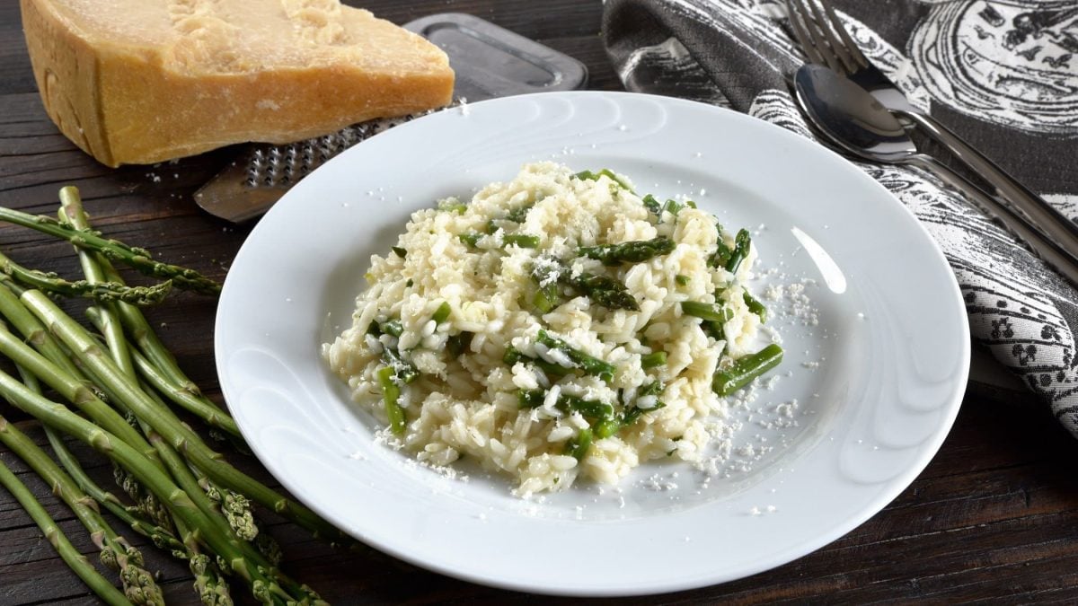 Draufsicht: ein Teller Risotto mit Wildspargel auf schwarzem Untergrund. Daneben liegen Wildspargel-Stangen, Parmesan und Besteck.