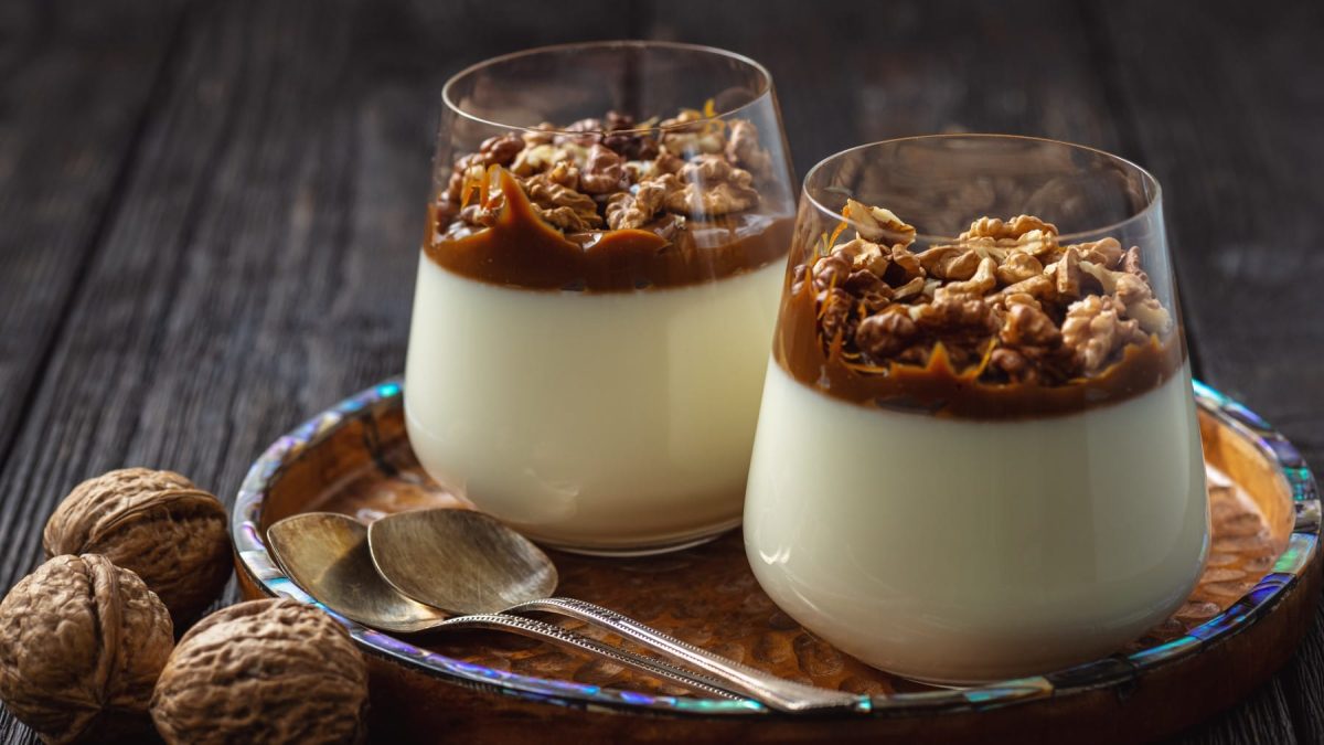 Zwei Gläser Sahnepudding mit Dulce de Leche und Walnüssen auf einem Tablett, daneben Löffel und Walnüsse vor Holzhintergrund.