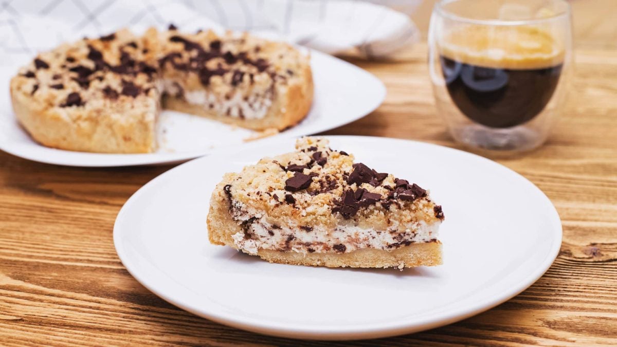 Ein Stück Sbriciolata alla Ricotta auf weißem Teller auf Holztisch. Im Hintergrund restlicher Kuchen und eine Tasse Espresso. Draufsicht.