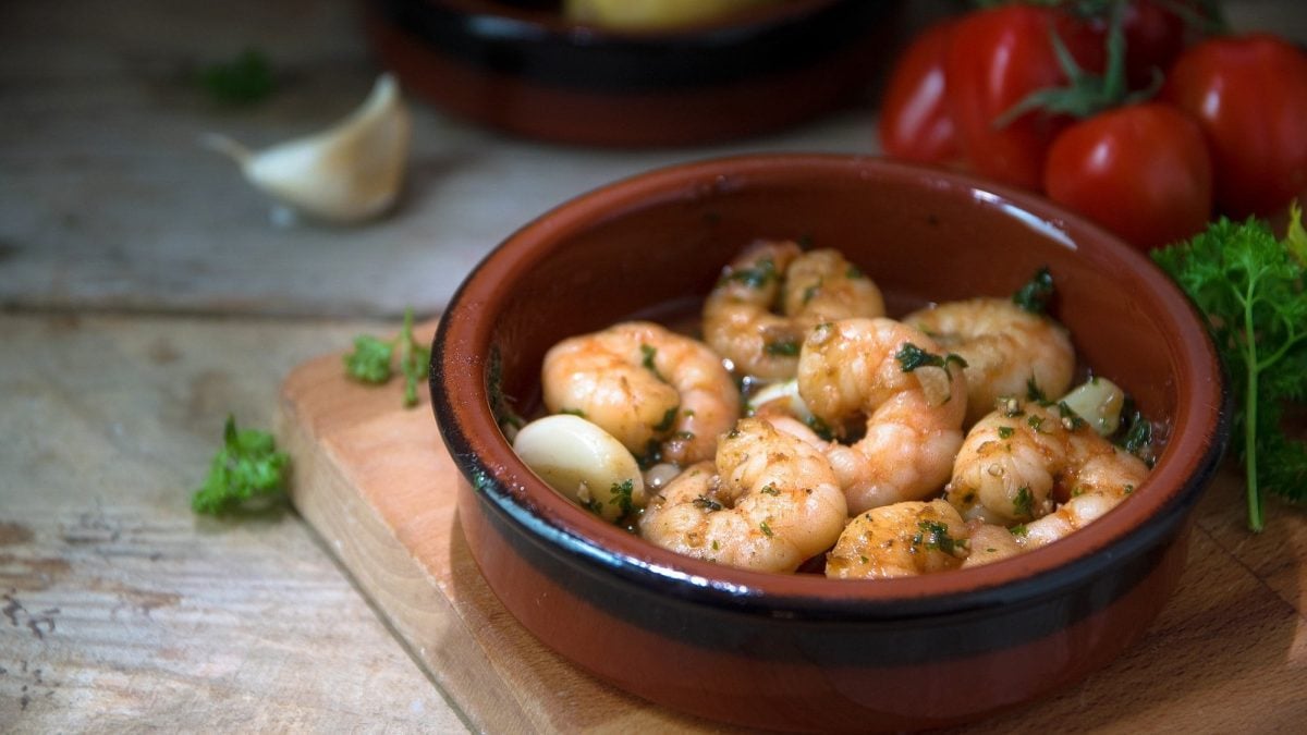 Frontalsicht: Gebratene Shrimps mit Knoblauch und Petersilie in Öl in einer Tonschüssel. Im Hintergrund sind rote Chilis und Knoblauchzehen.