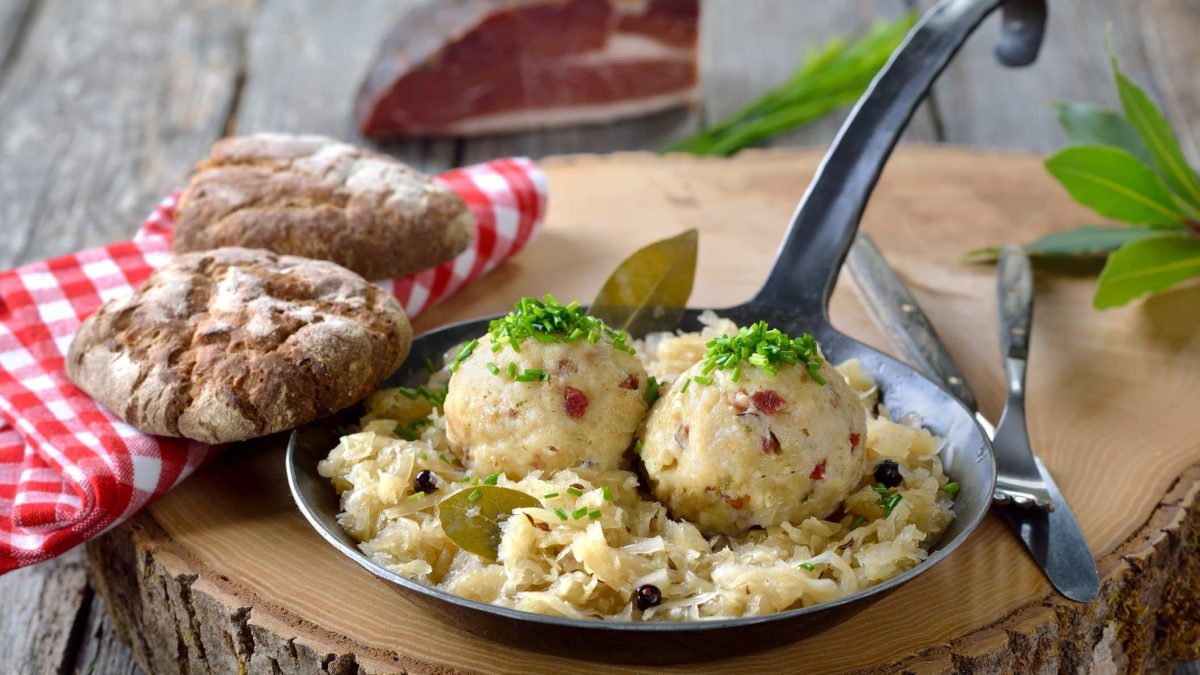 Frontalsicht: Auf einem Holztisch steht eine gusseiserne Pfanne Speckknödel mit Sauerkraut. Drum herum liegen Gewürze und ein weiß-rot kariertes Tuch.