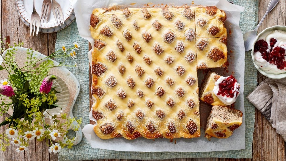 Angeschnittener Steppdecken-Mohnkuchen auf Backpapier vor hellem Hintergrund in der Draufsicht, daneben Blumen, Geschirr und Quark mit Beeren.