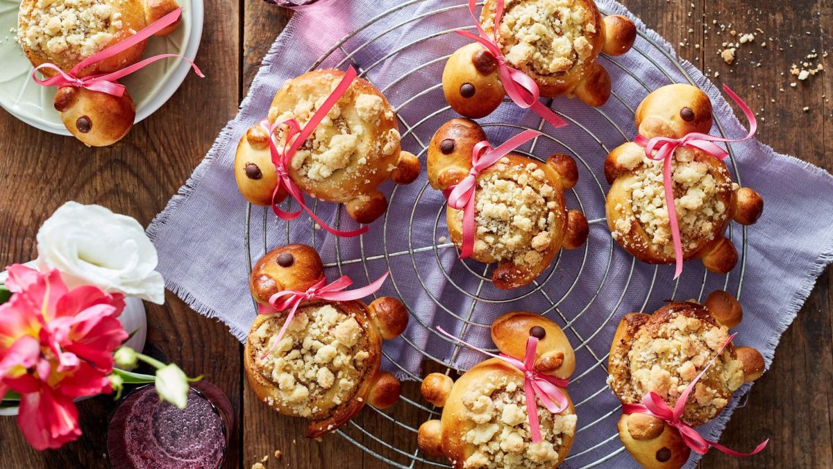 Mehrere Streusel-Lämmer mit rosa Schleifen auf einem Gitter vor lila Küchentuch in der Draufsicht.