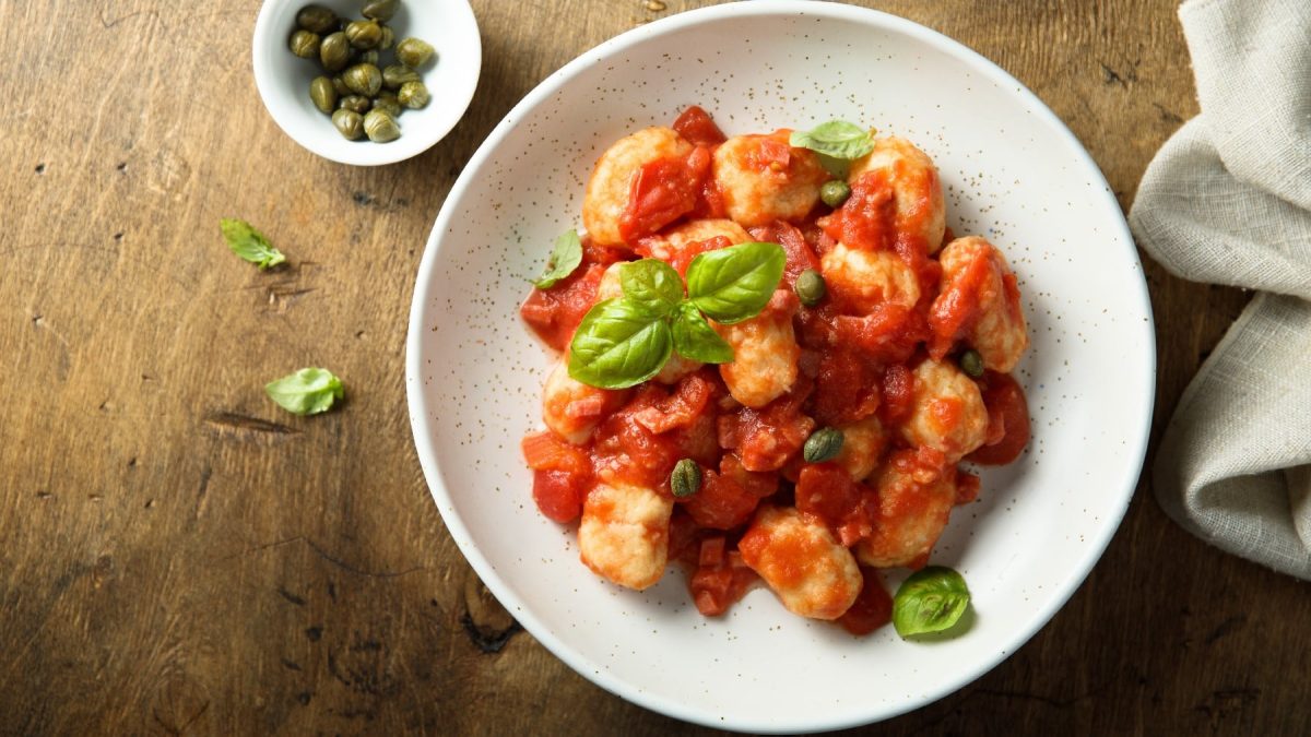 Ein weißer Teller mit den Mehl-Gnocchi und der Sucuk-Tomatensauce mit Kapern auf einem Holztisch.