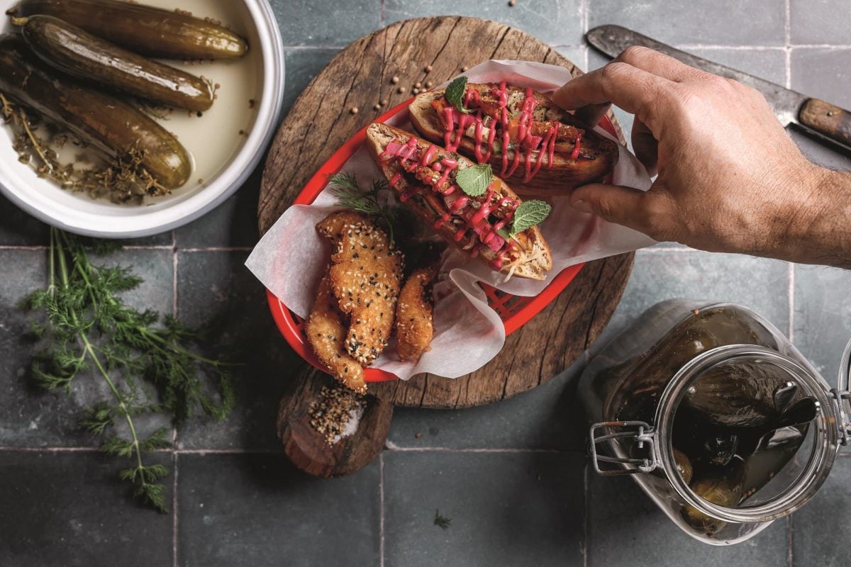 Sum Sum Schnitzelim, kleine Schnitzel in geröstetem Zatar-Pita-Brot, nach denen eine Hand greift. Daneben eine Schüssel und ein Glas Gewürzgurken. Alles Draufsicht.