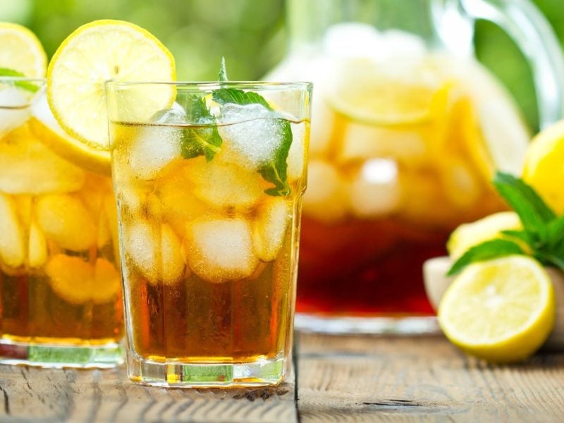 Zwei Longdrink-Gläser vor einer Karaffe mit Zitronentee auf einem Holztisch.