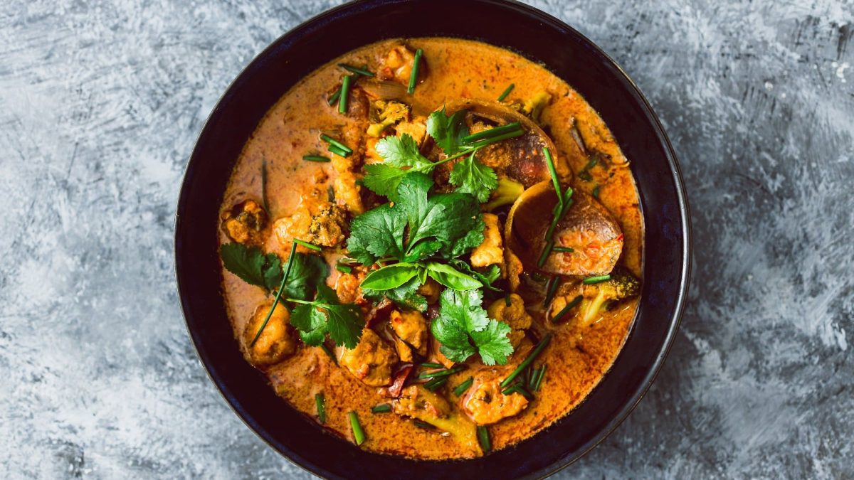 Draufsicht: Auf einem grauen Untergrund steht eine schwarze Schüssel Tofu-Curry mit Brokkoli und Koriander.
